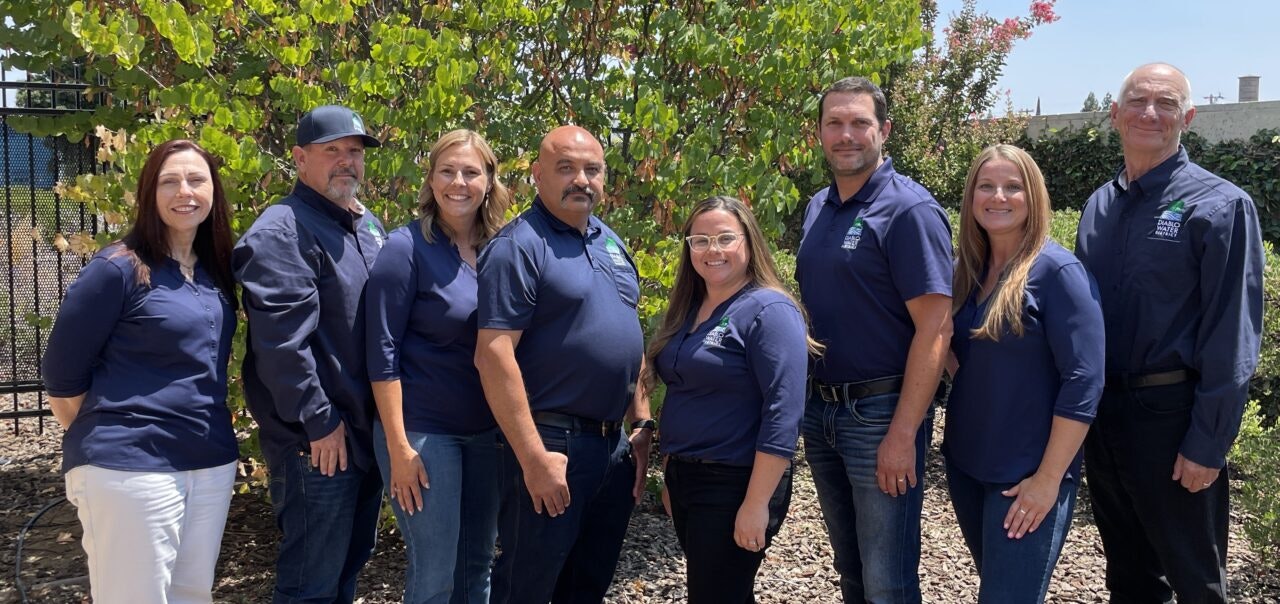 A group of eight people in matching blue shirts posing outdoors in front of greenery.
