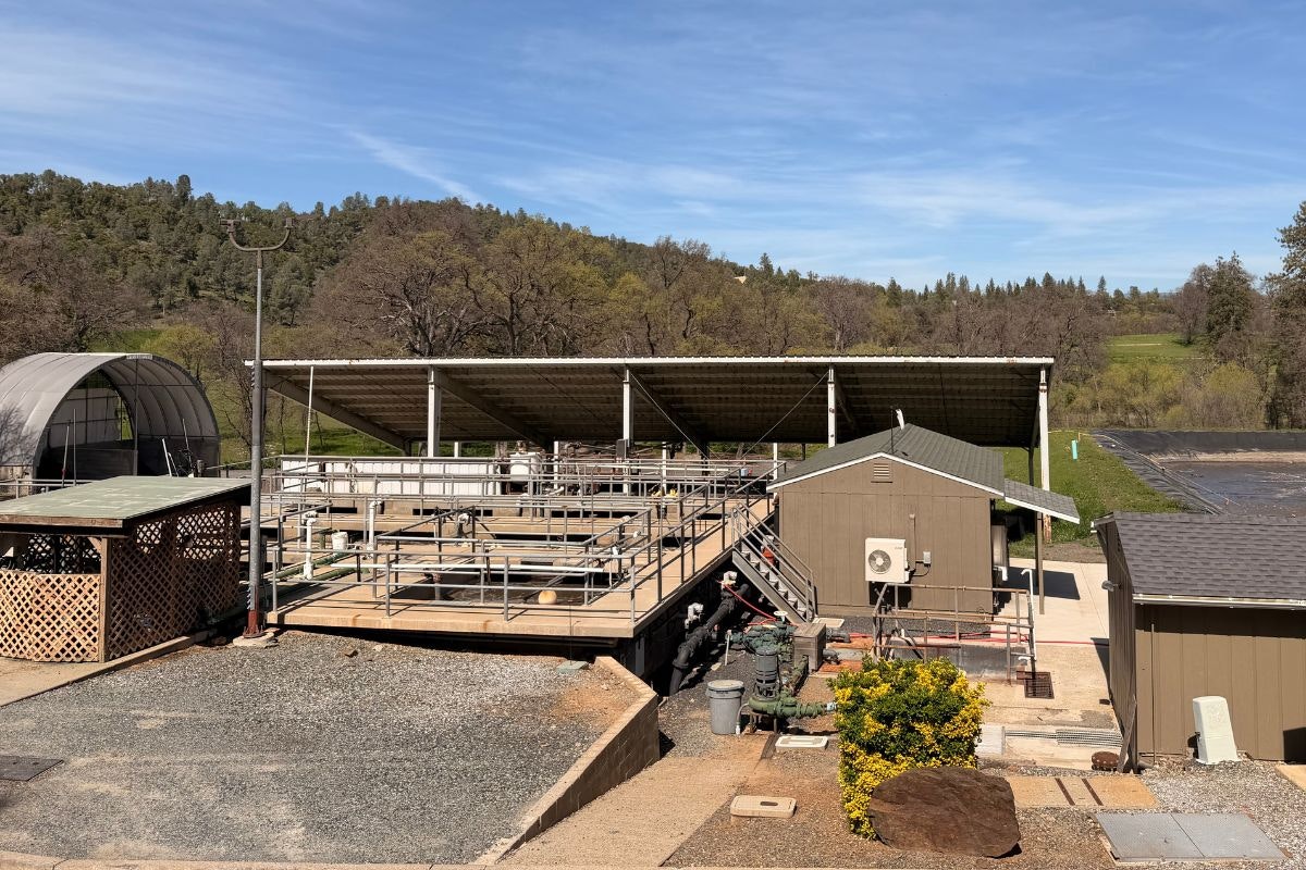 The image shows a rooftop structure surrounded by buildings, equipment, and a natural landscape with trees and hills in the background.
