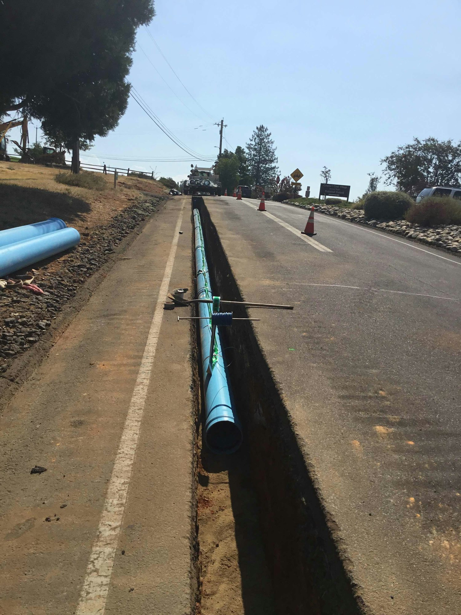 A blue pipe is being installed in a trench along a roadside, with orange traffic cones guiding vehicles.
