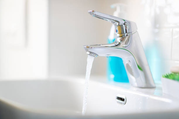 A modern chrome faucet with running water in a white sink; a blue soap dispenser is in the background.