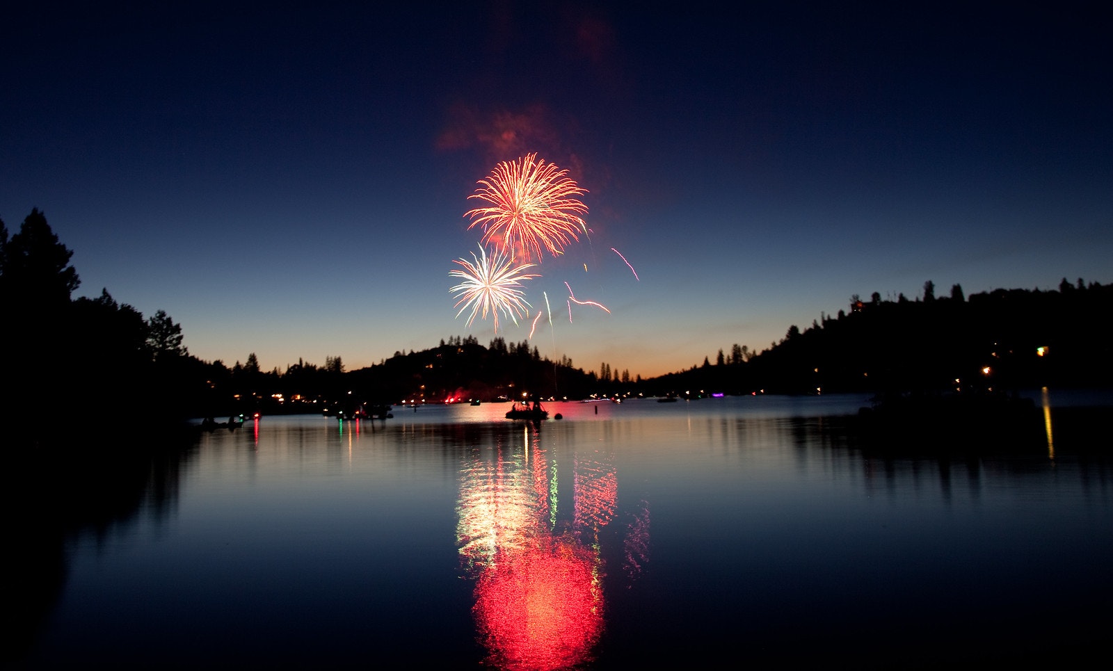 Fireworks over a serene lake at dusk with trees silhouetted against the twilight sky and reflections on the water.