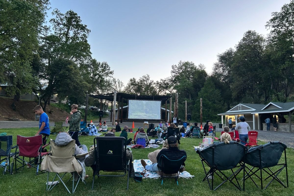 A crowd sits on lawn chairs watching a movie on a large screen in an outdoor setting surrounded by trees.
