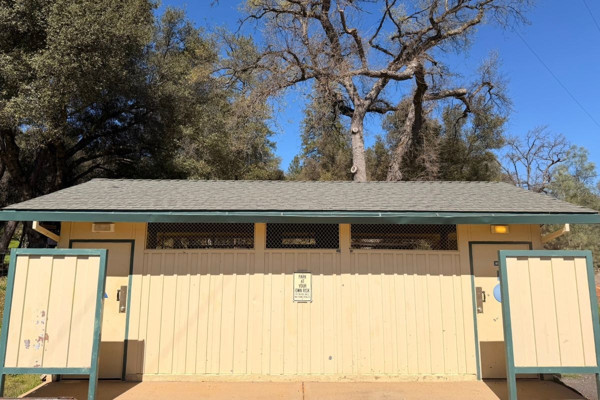 The image shows a small restroom building with two doors, surrounded by trees and a clear blue sky.