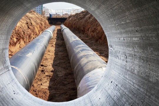 Two large pipes in a trench viewed through a concrete tunnel opening, with construction in the background.