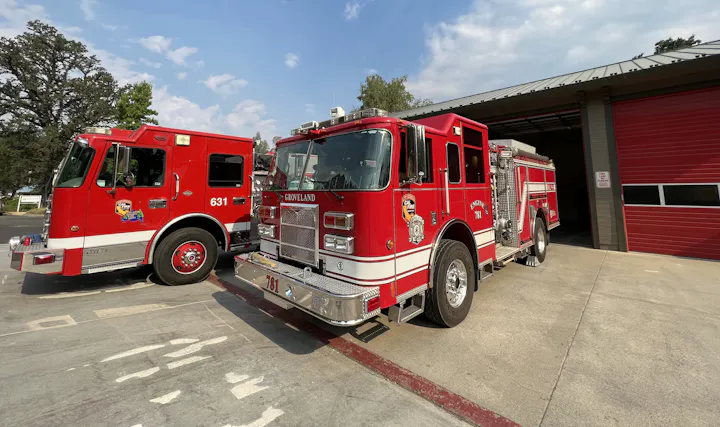 Two red fire trucks parked outside a fire station with open doors.