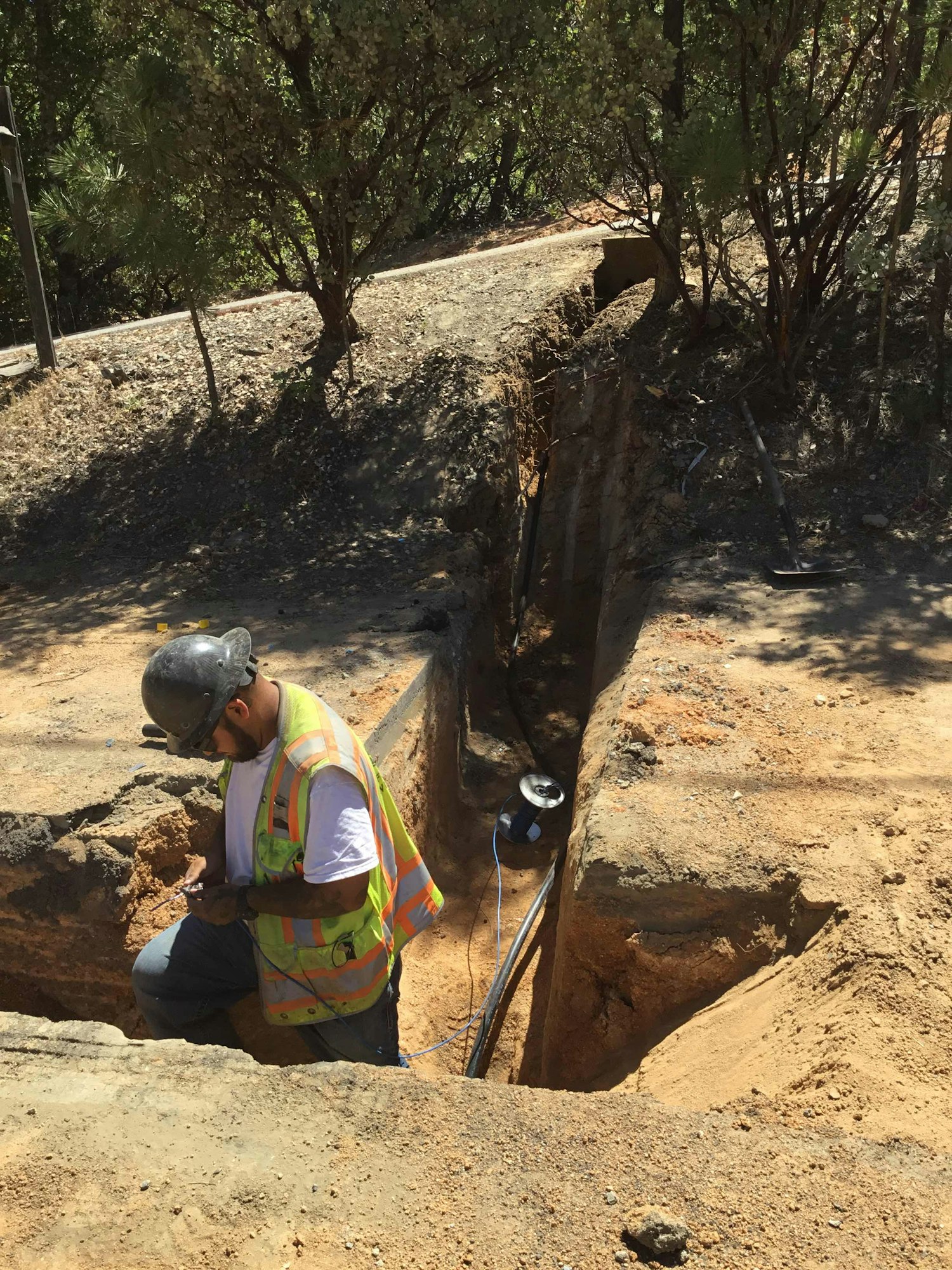 A construction worker in a safety vest and helmet is standing in a trench with tools and wiring nearby, surrounded by trees.