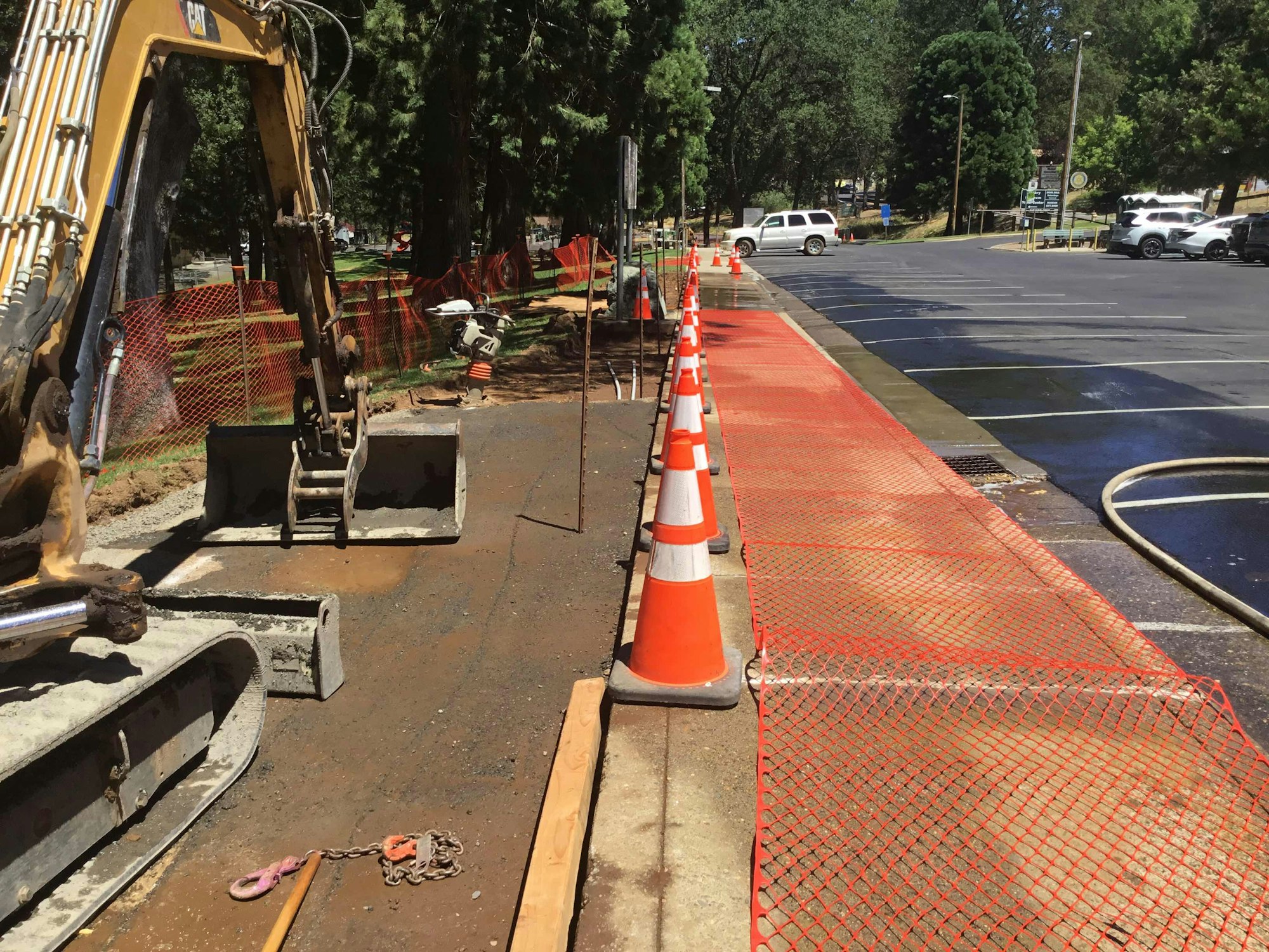 Construction site with an excavator, orange cones, and netting along a sidewalk near a parking lot.