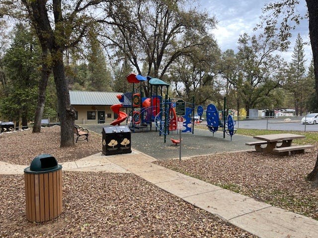Playground with slides and climbing structures, surrounded by trees; benches, picnic table, garbage bin, and a building in the background.