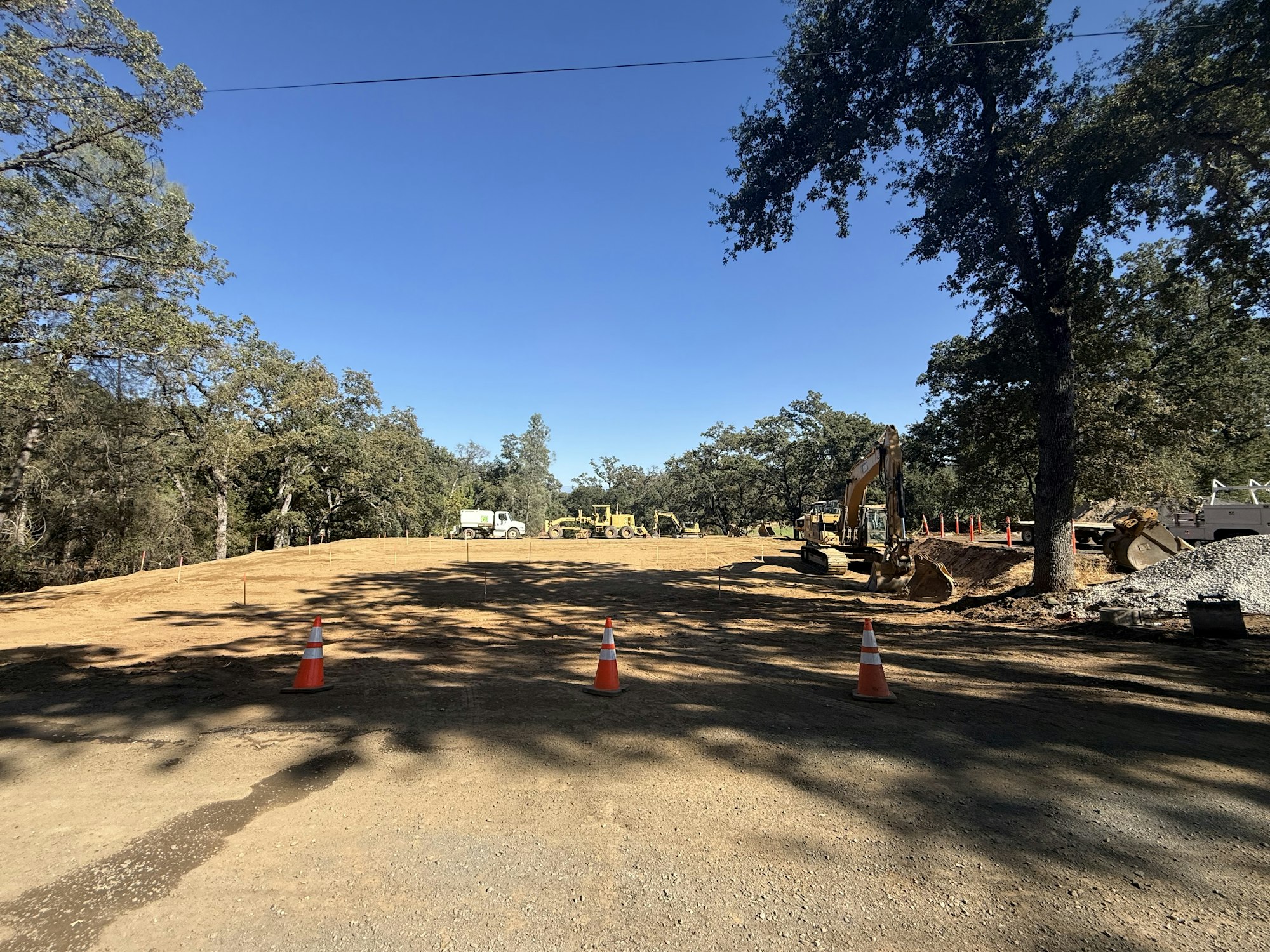 Construction site with machinery, traffic cones, dirt area, and trees under a clear blue sky.