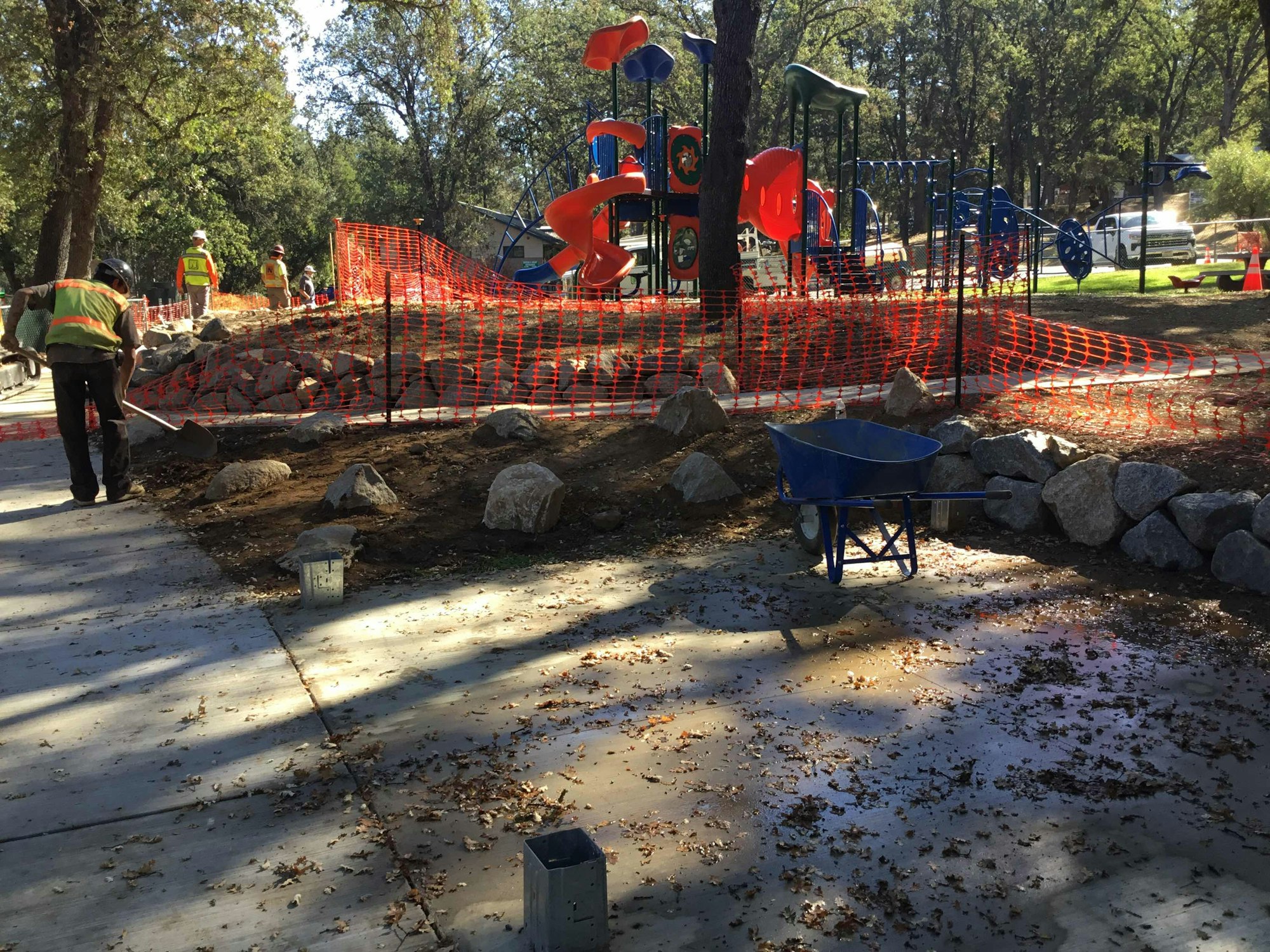 Construction site with workers near a playground, orange safety fencing, wheelbarrow, and rocks.