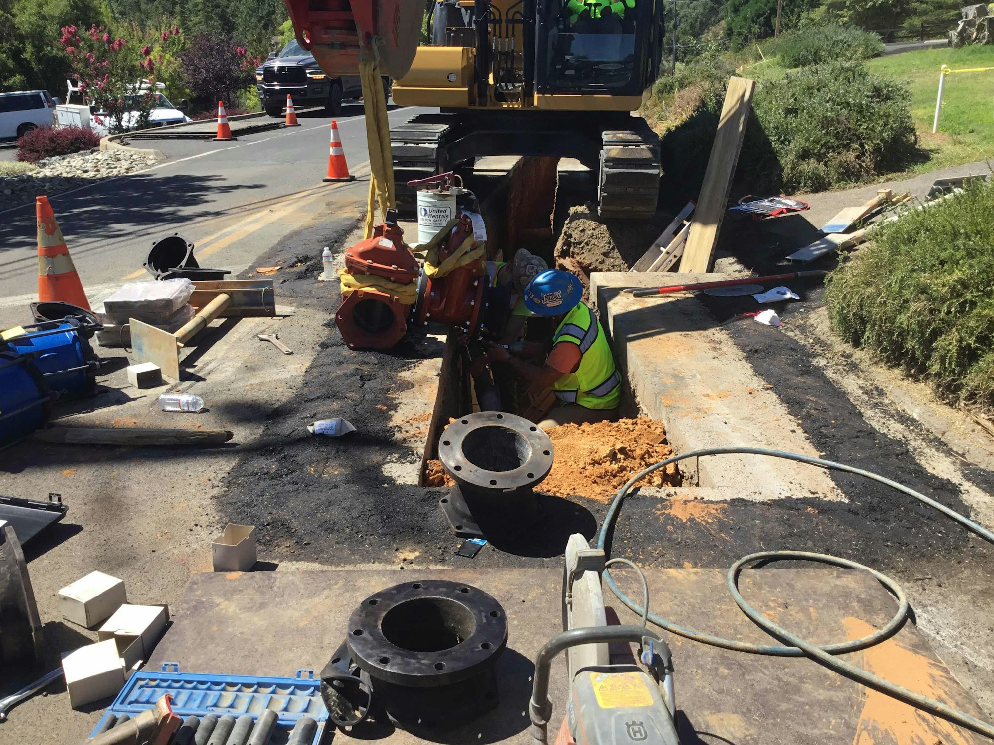 A construction worker in a trench, surrounded by machinery and equipment, works on a road with traffic cones nearby.