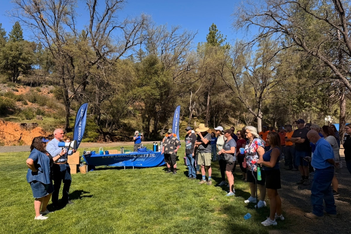 A group of people gather outdoors, listening to speakers with a table displaying items, under clear skies and trees.