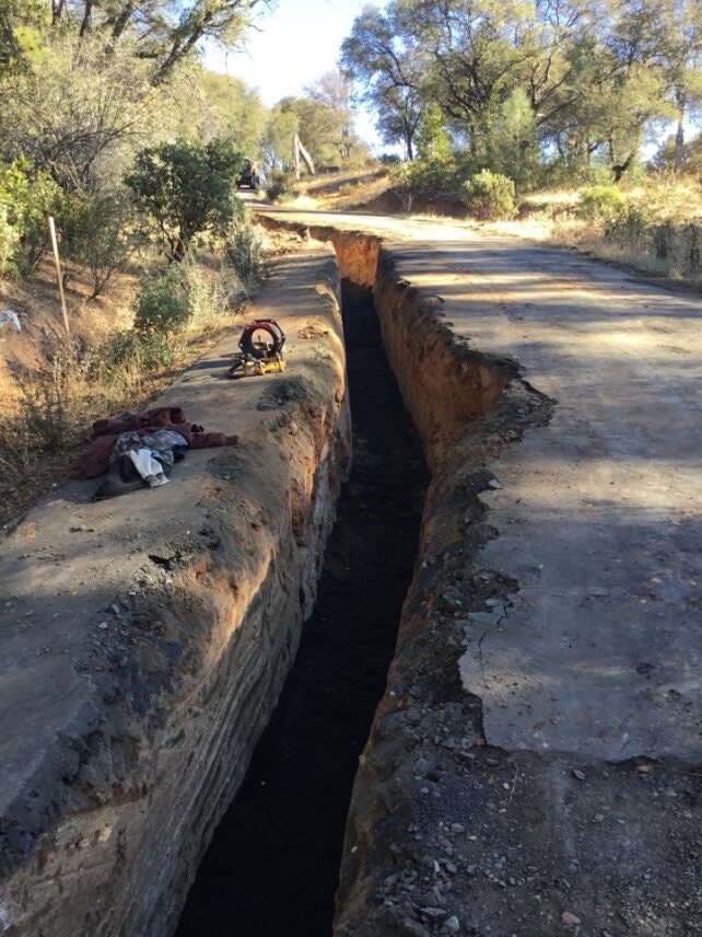 A narrow trench dug alongside a rural dirt road with some tools and clothes lying nearby.