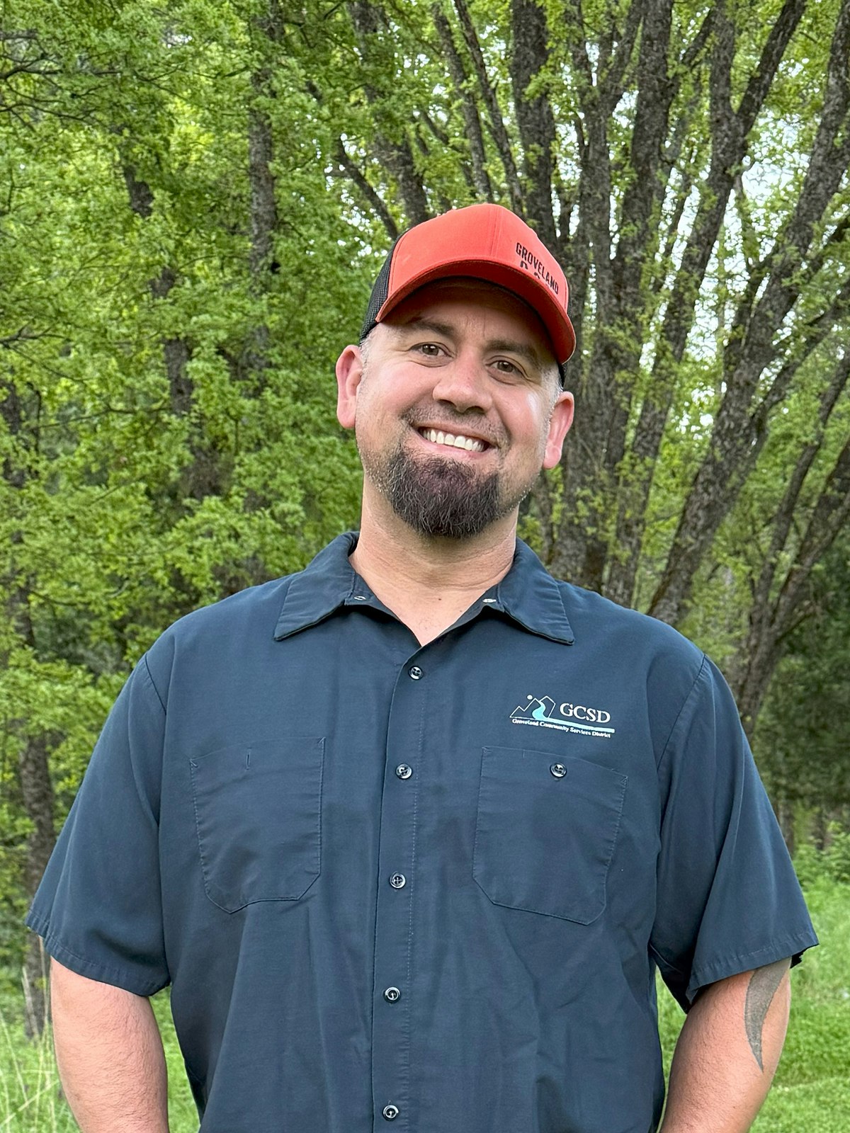 A person in a blue shirt and red cap standing outdoors in front of green trees, smiling.