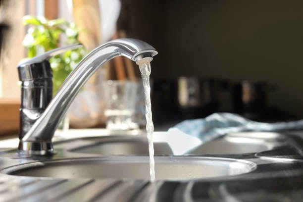 A kitchen faucet with running water over a sink, a glass, and potted plant visible in the background.