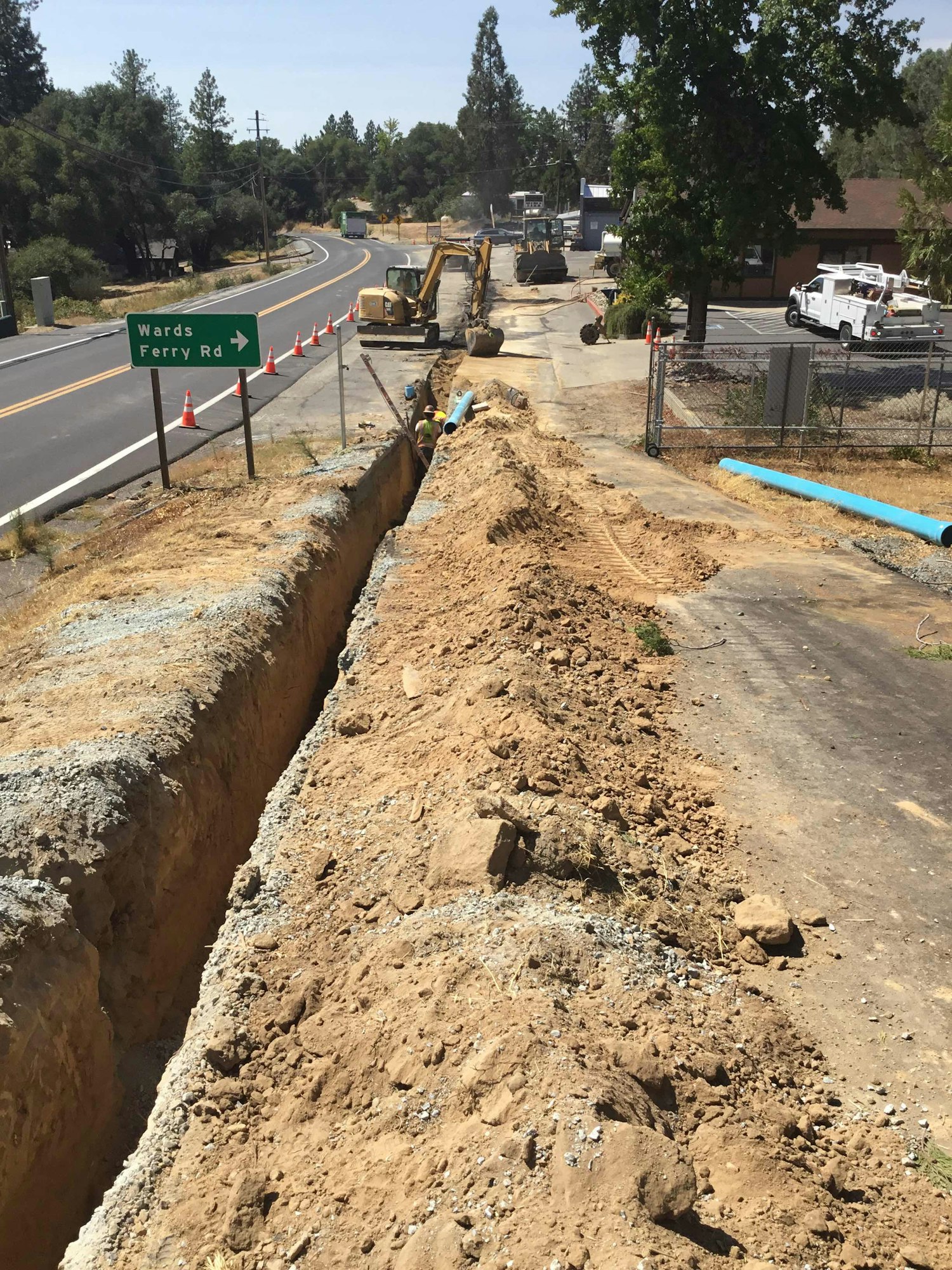 Roadside construction with excavators, cones, and workers laying blue pipes near Wards Ferry Rd.