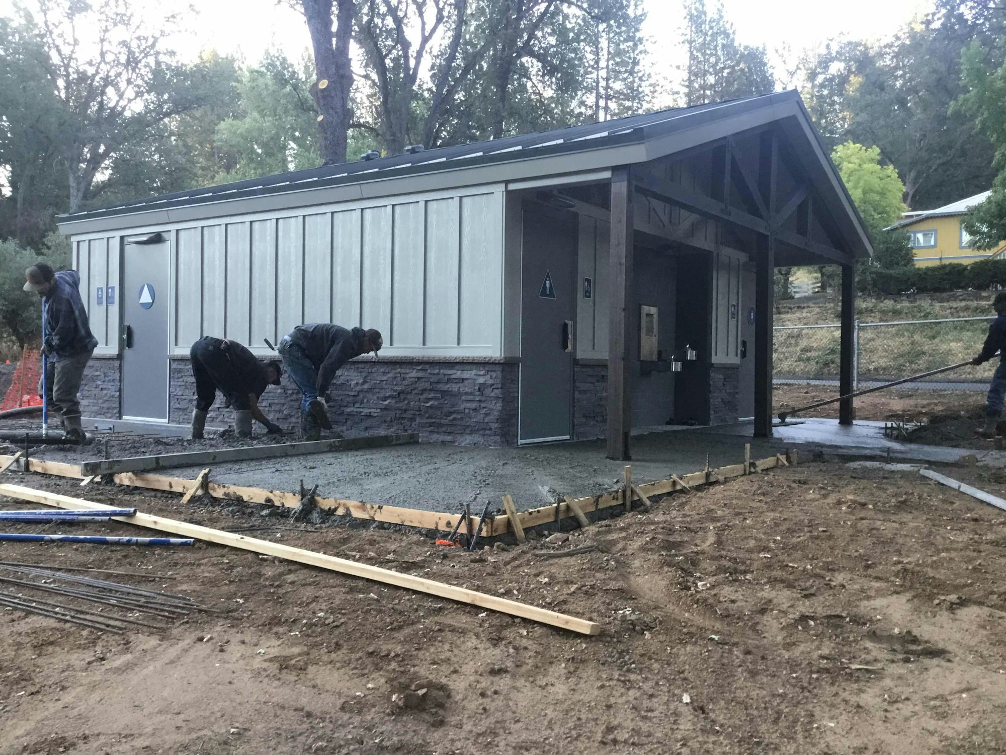 Workers are laying concrete outside a small building surrounded by trees.
