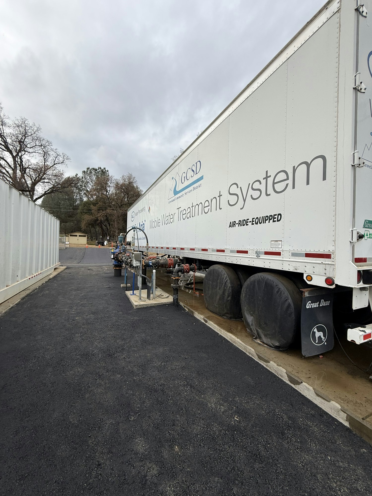 The image shows a large trailer labeled "Waste Water Treatment System" parked alongside equipment and a gravel area, under cloudy skies.