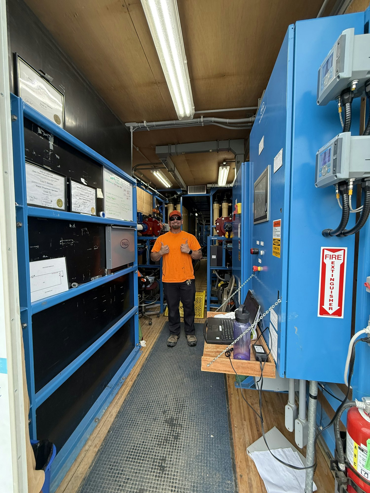 The image shows a workspace with blue machinery, a person in an orange shirt, and various equipment and safety signs.