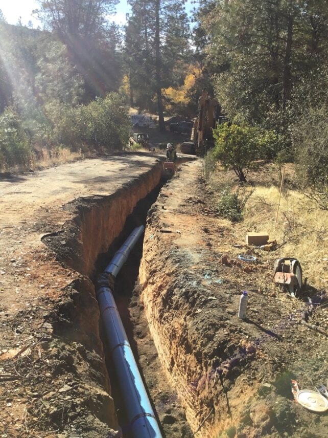 Construction site with a trench and installed pipeline, surrounded by trees and equipment.