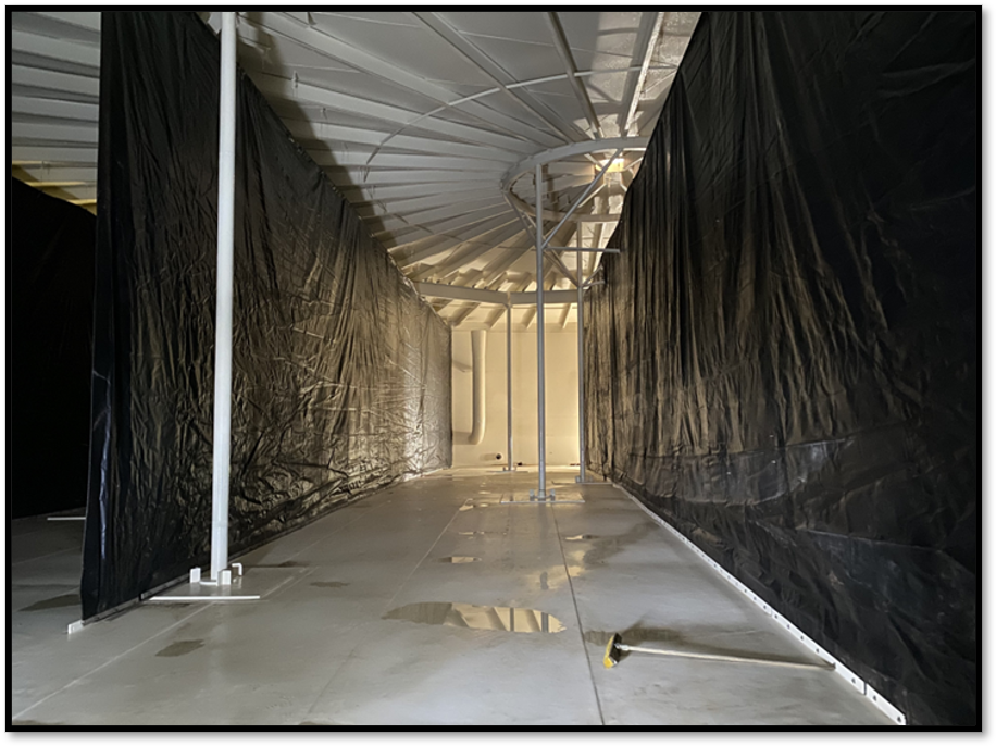 Interior view of a metal structure with black plastic sheeting, some poles, and a wrench on the floor.