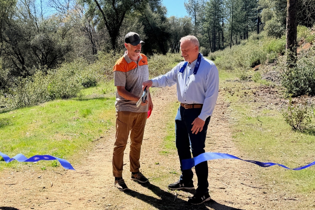 Two men are smiling and engaging in a ribbon-cutting ceremony in a wooded area, celebrating an event or opening.