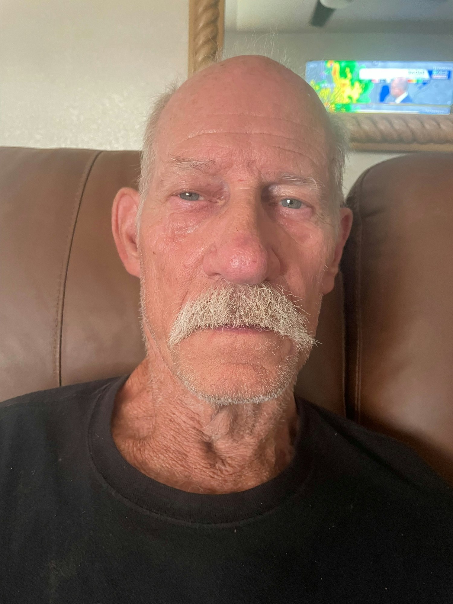 A man with a mustache is sitting on a couch, looking directly at the camera, with a TV showing news in the background.