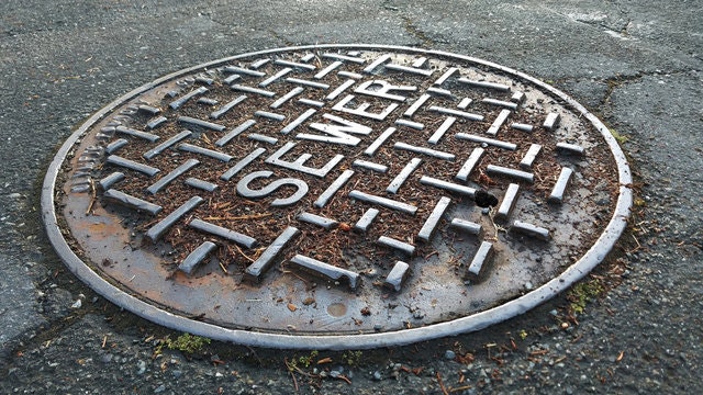 Round manhole cover labeled "SEWER" with a checkered pattern, surrounded by a concrete surface.