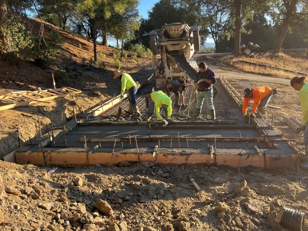 Workers pouring and leveling concrete on a construction site with a mixer truck and wooden forms visible.
