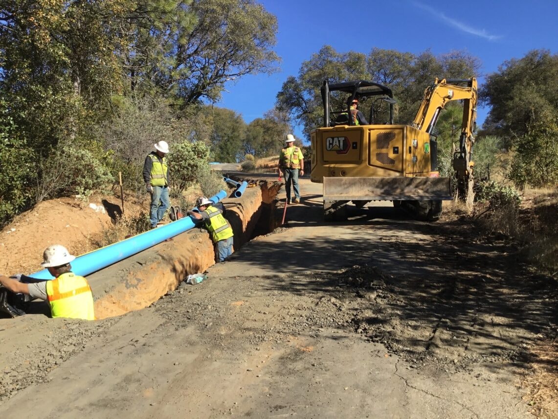 Workers installing a large blue pipe in a trench with a backhoe nearby, surrounded by trees and a clear blue sky.