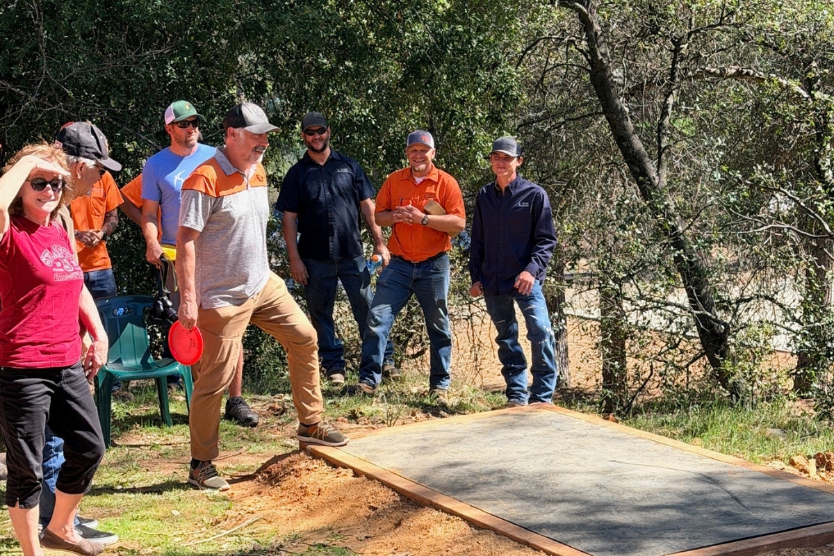 A group of people are gathered outdoors, watching someone about to throw a frisbee or disc from a wooden platform.