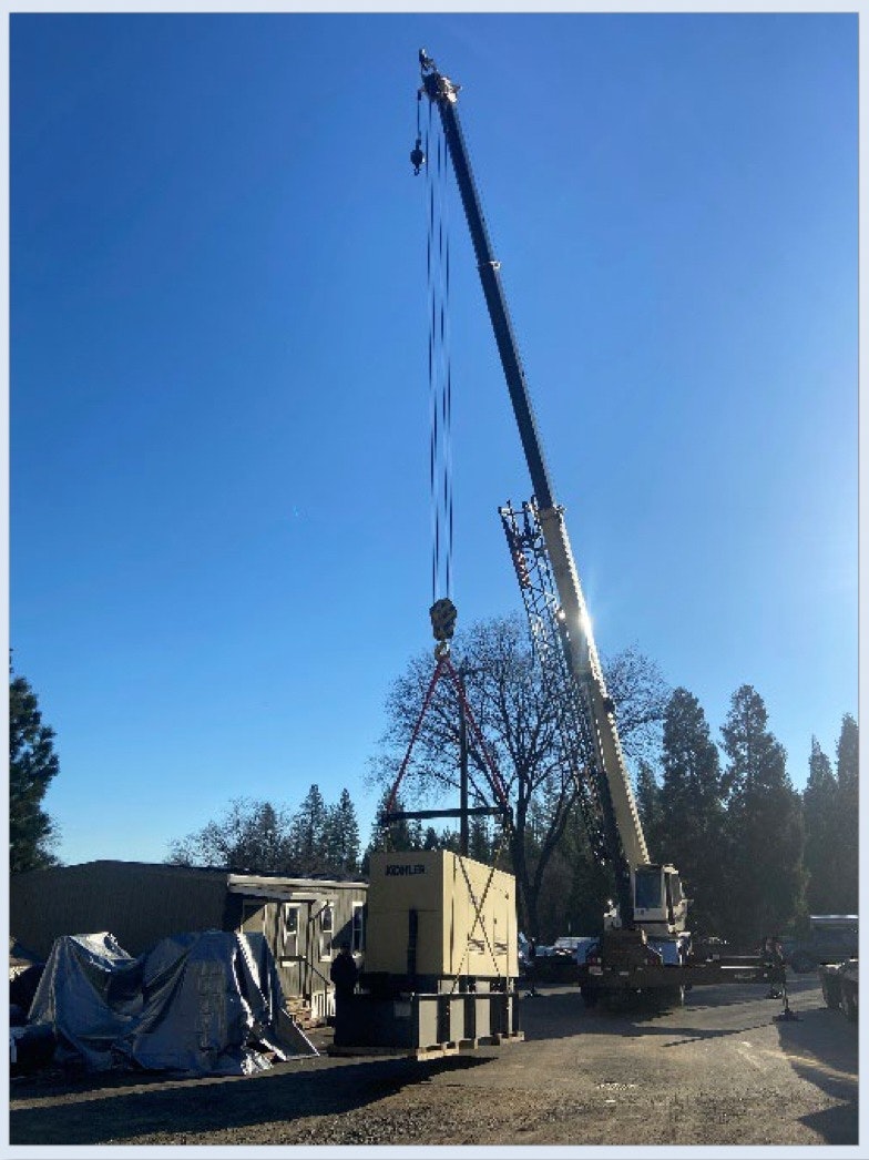 A crane is lifting a large generator near a building under a clear blue sky. Trees are visible in the background.