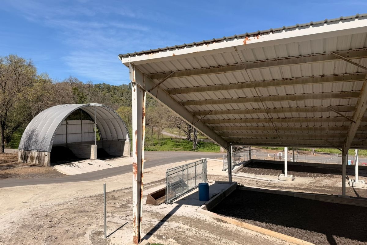 The image shows two large metal canopies at a rural site, with a dirt area, fencing, and a clear blue sky in the background.