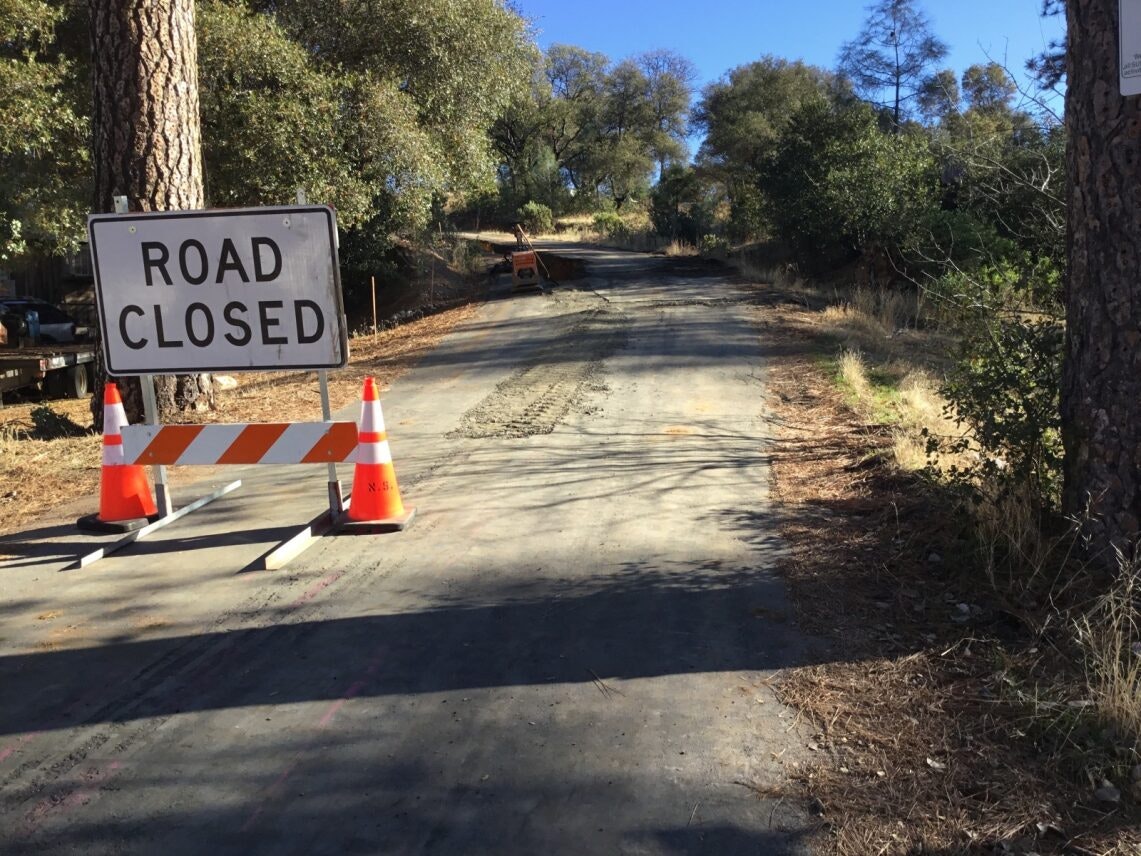 A "Road Closed" sign with traffic cones blocks a rural road amid trees.