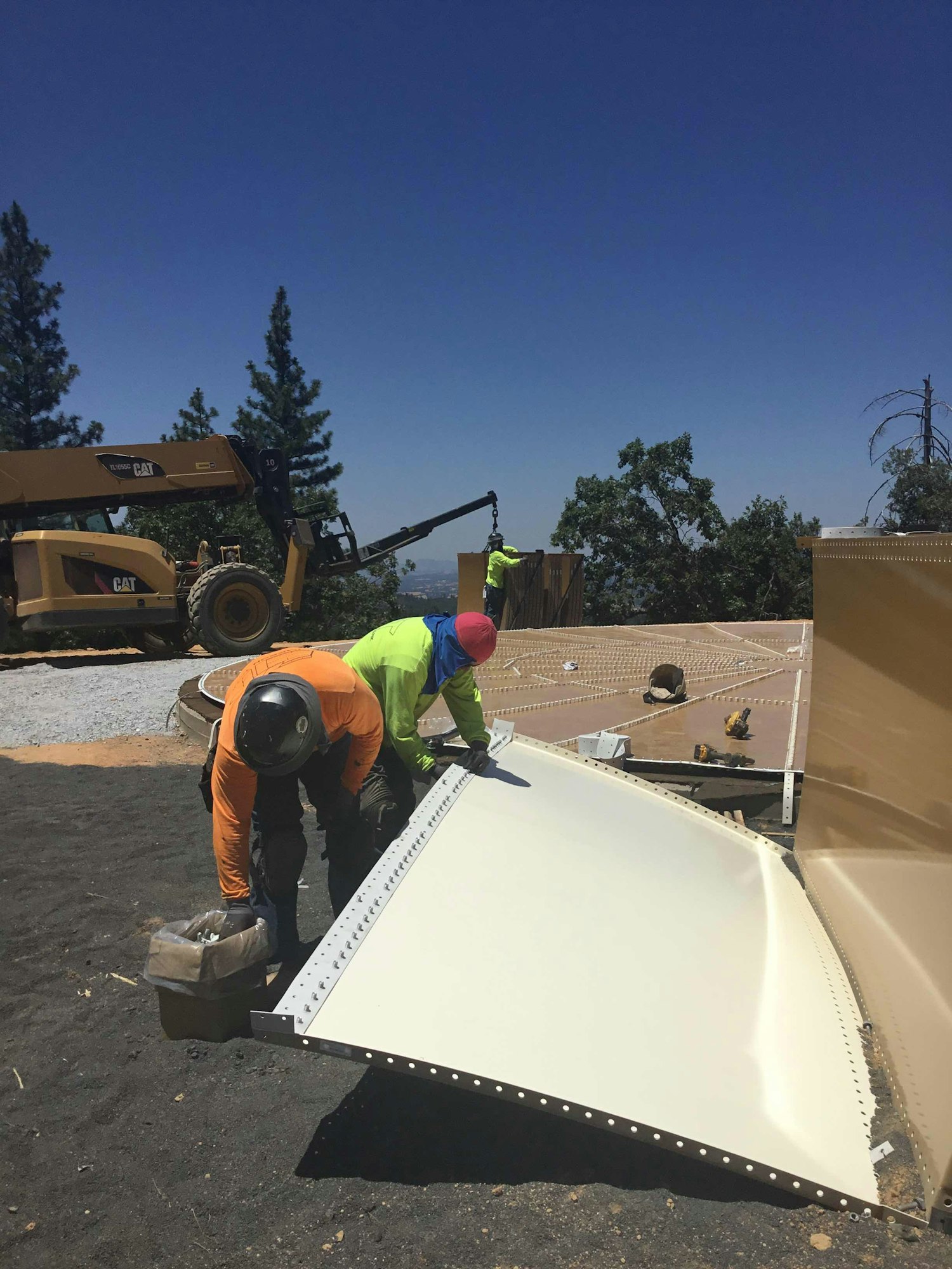 Two workers in safety gear handling a large metal panel at a construction site with a crane and trees in the background.