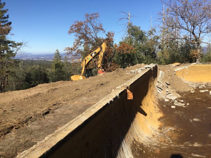 Excavator near a cleared dirt area with broken concrete, surrounded by trees and hills in the background.