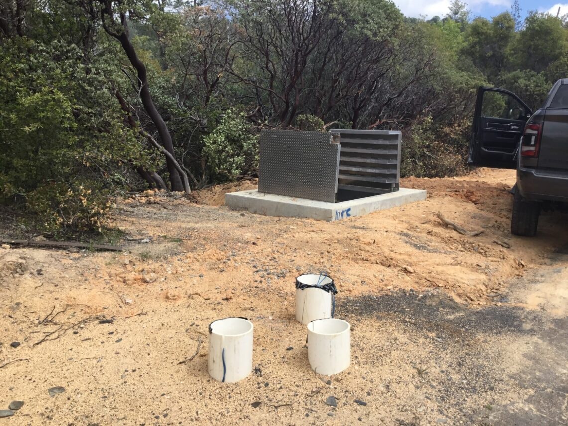 Outdoor scene with a truck, a metallic structure on a concrete base, and three white buckets on the ground near trees.