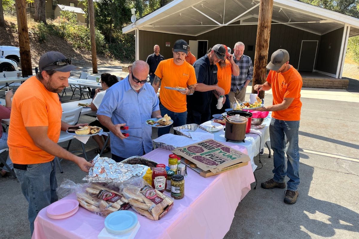 A group of people are enjoying a meal outdoors, serving food from a table with various dishes and drinks available.