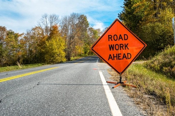 Orange "ROAD WORK AHEAD" sign by a rural road surrounded by trees.