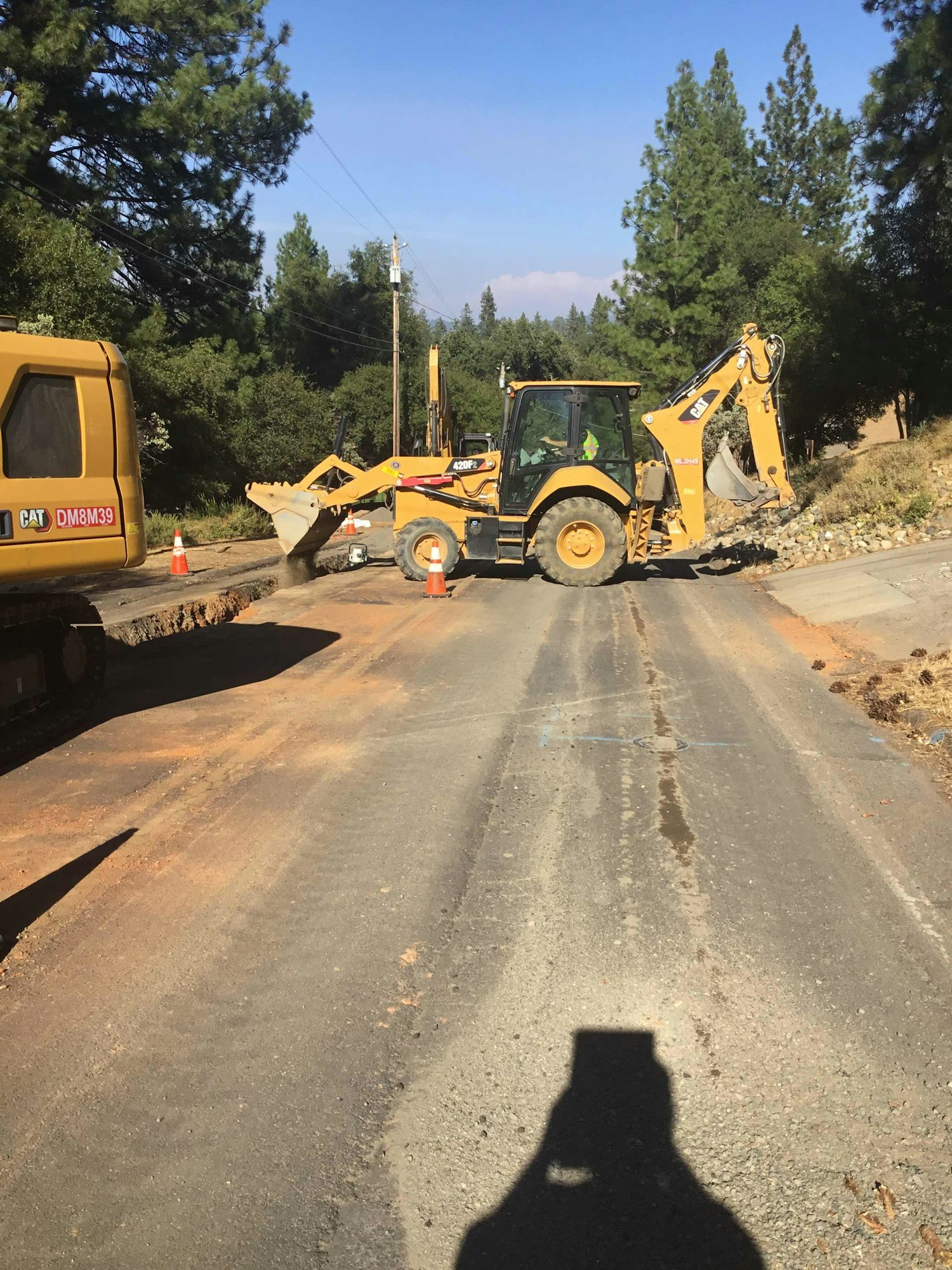 Roadwork with a backhoe loader and construction equipment on a rural road, surrounded by trees and traffic cones.