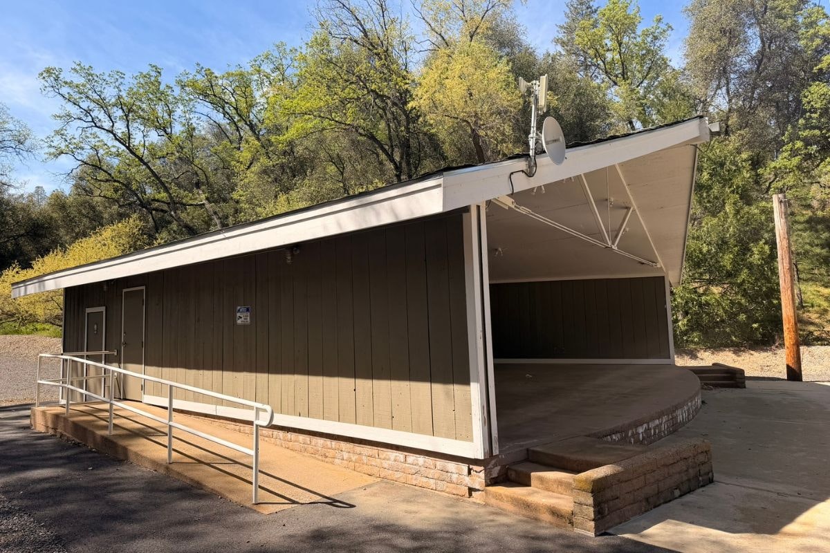 A small building with a ramp, partially covered porch, and trees in the background on a sunny day.