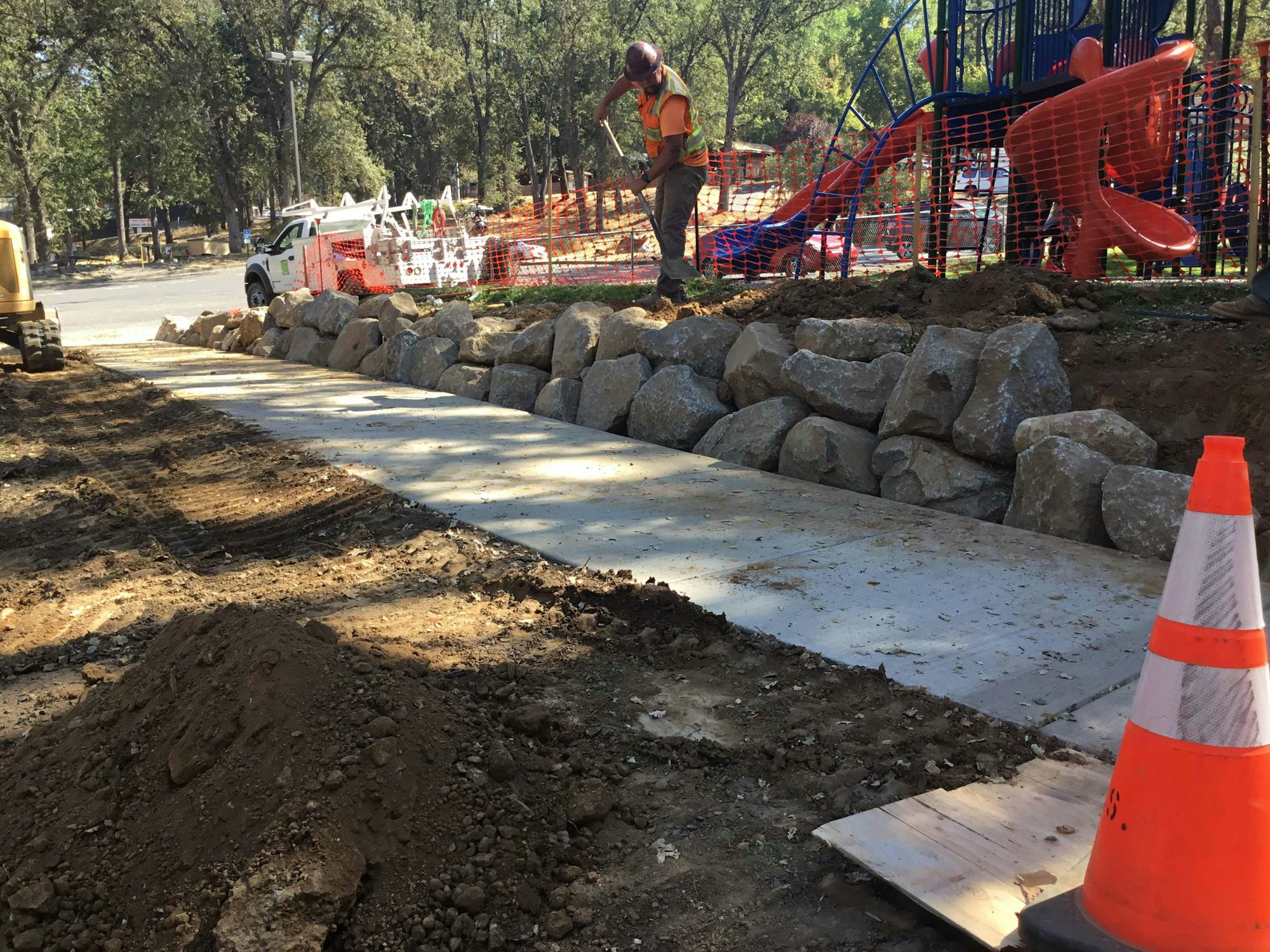 Construction site with a sidewalk, rocks, and playground; worker in a safety vest using a hoe; orange cones for caution.