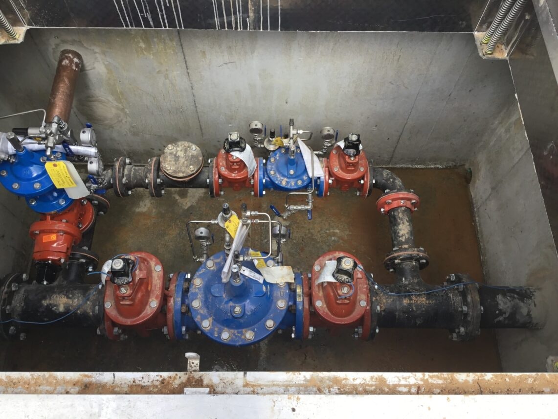A complex arrangement of industrial valves, pipes, and connectors in a concrete enclosure, with blue and red components.