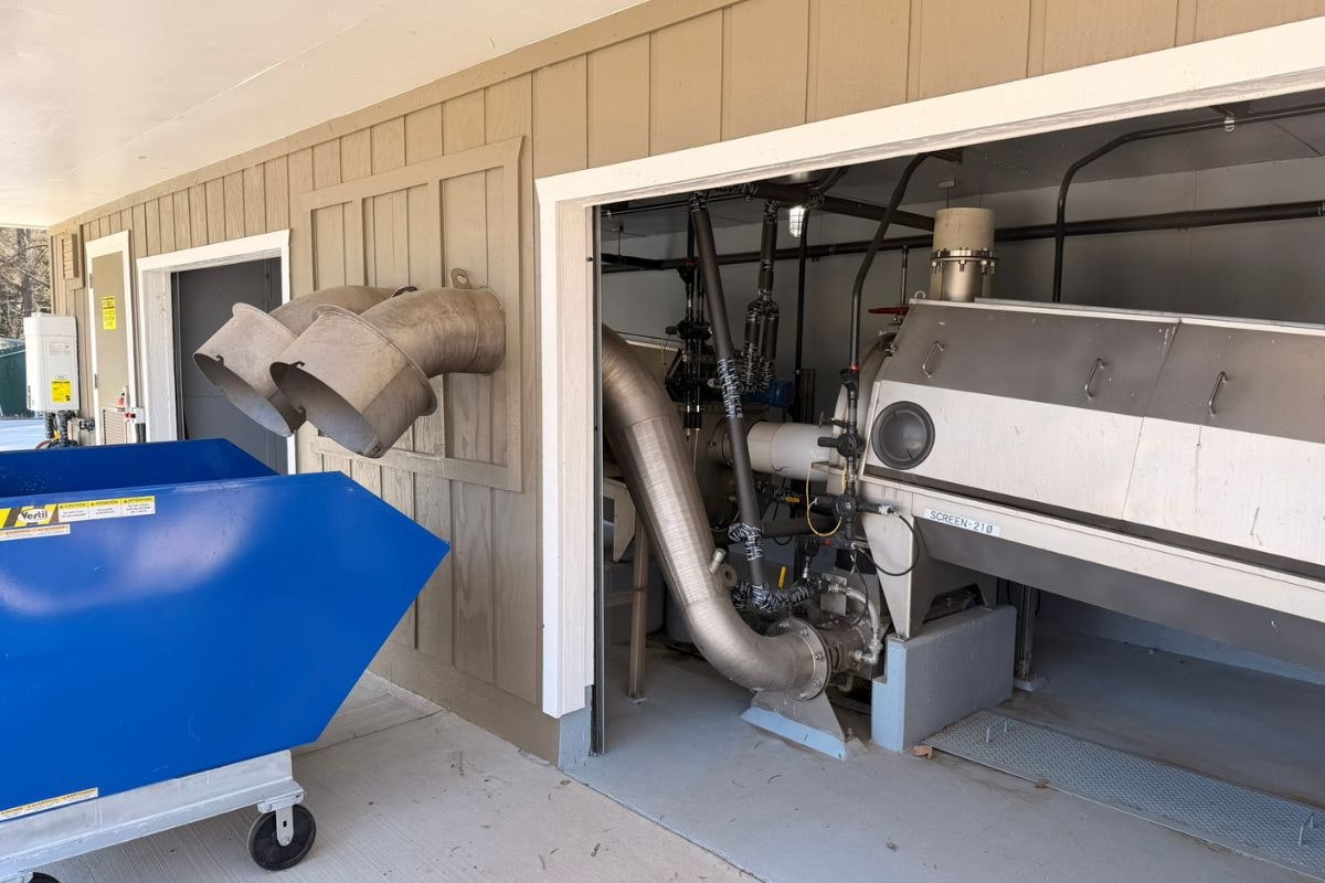 The image shows a mechanical setup with large pipes and equipment inside a building, alongside a blue container outside.