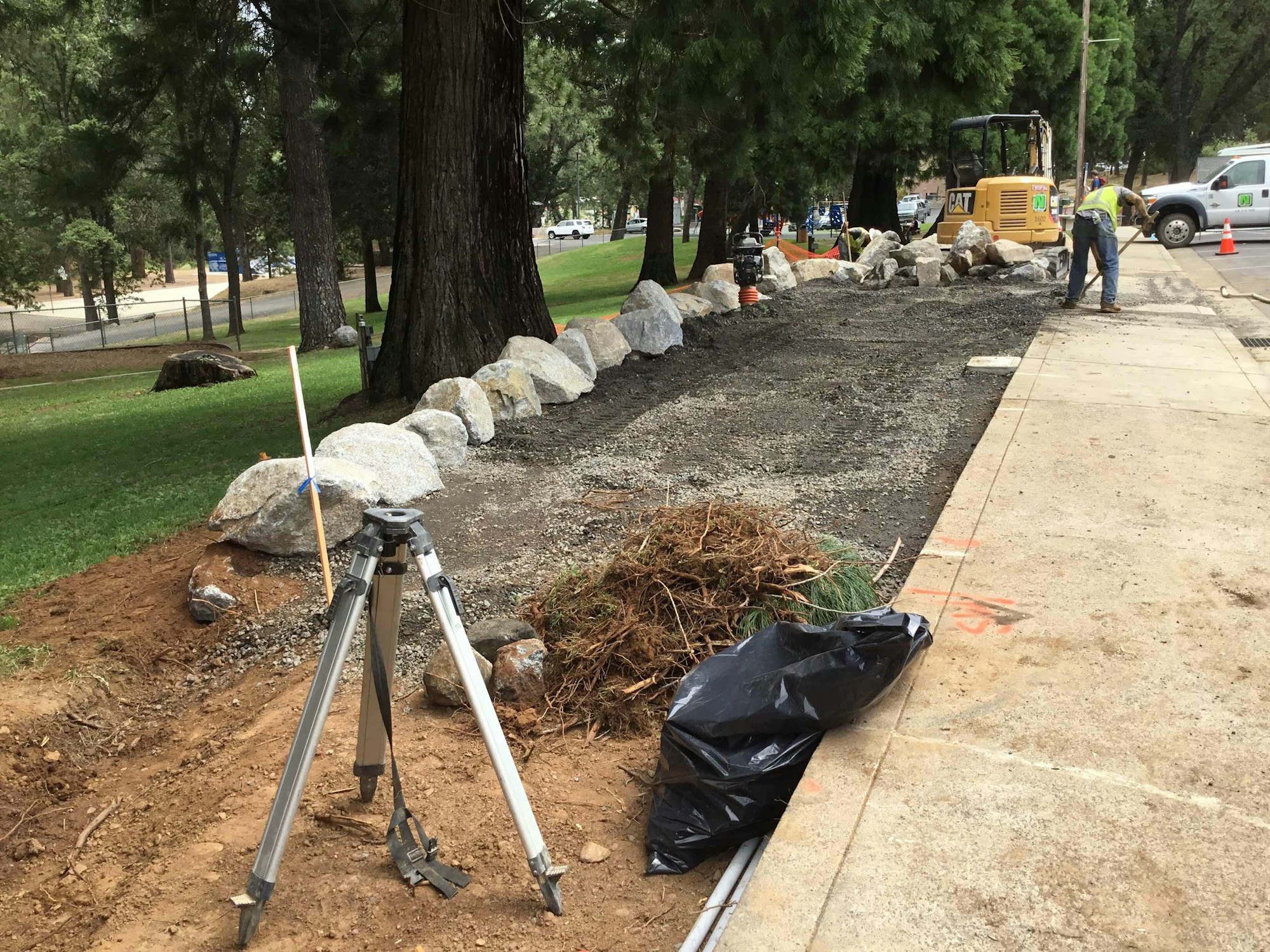 Construction site with heavy machinery, tripod, rocks, and a path being built beside trees.