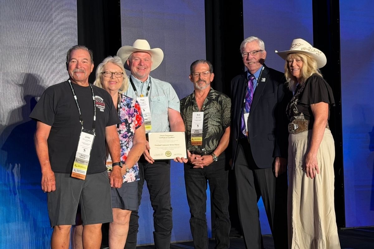 A group of six individuals stands on stage, holding a certificate, likely at an awards or recognition event.