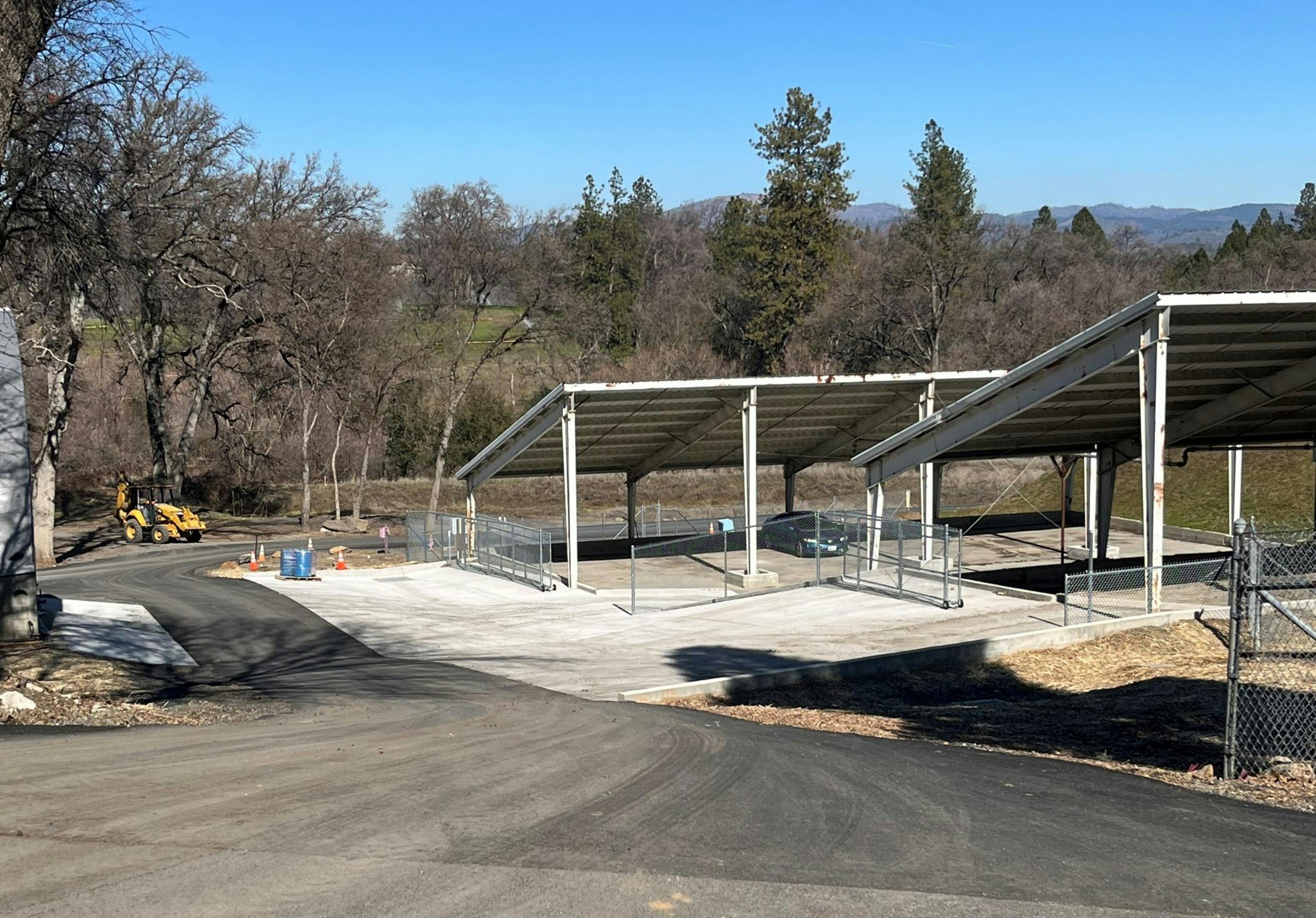 The image shows a construction site with partially finished covered structures, a dirt road, and some equipment in an outdoor setting.