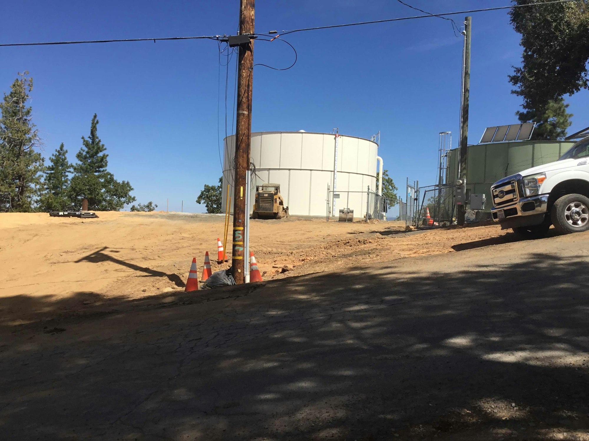 A construction site with large tanks, cones, a truck, and trees under a clear blue sky.