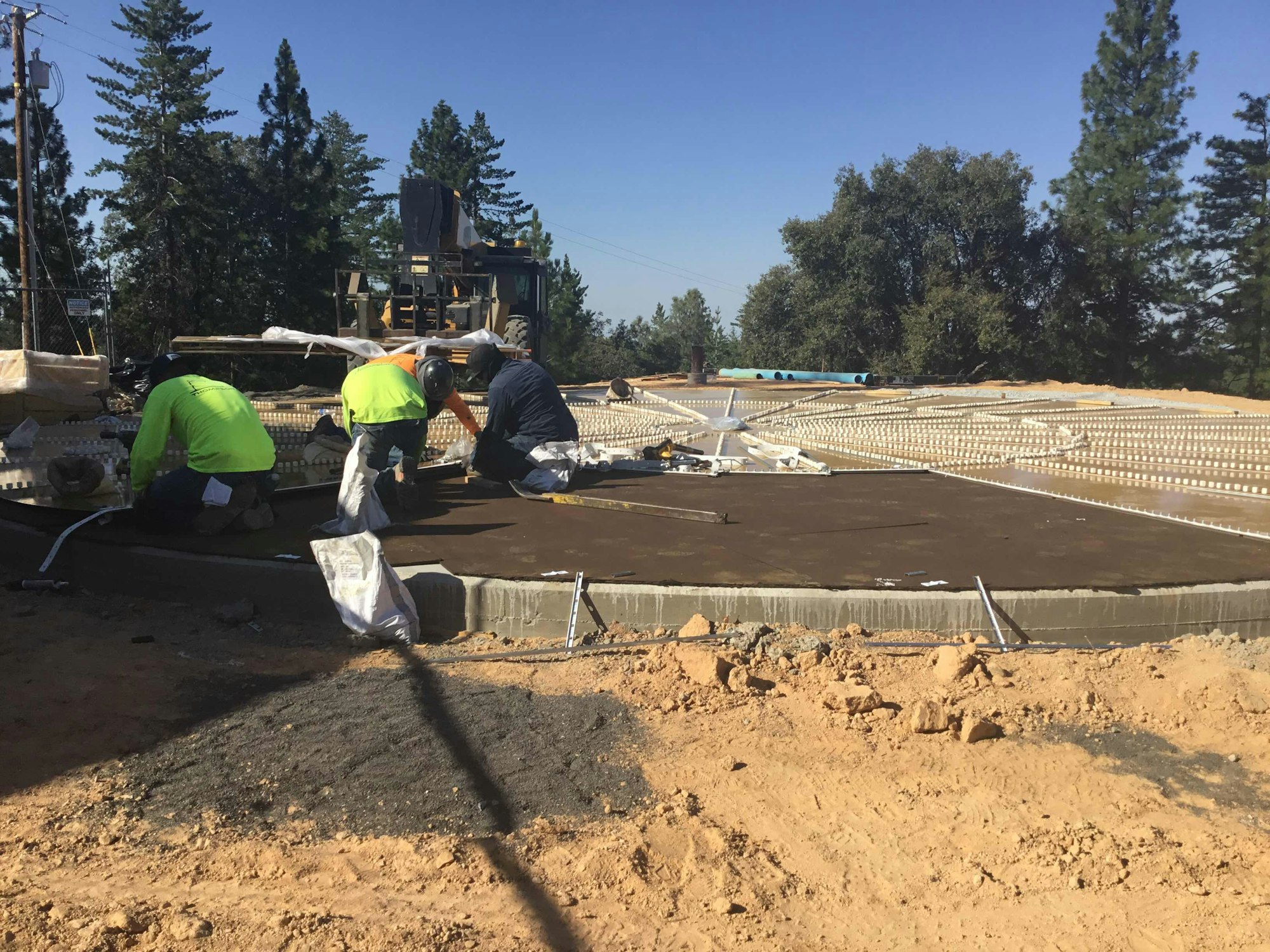 Three construction workers in safety gear working on a concrete foundation at a construction site with machinery and trees in the background.