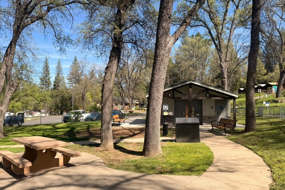 A park scene with trees, a picnic table, benches, and a restroom facility on a sunny day.